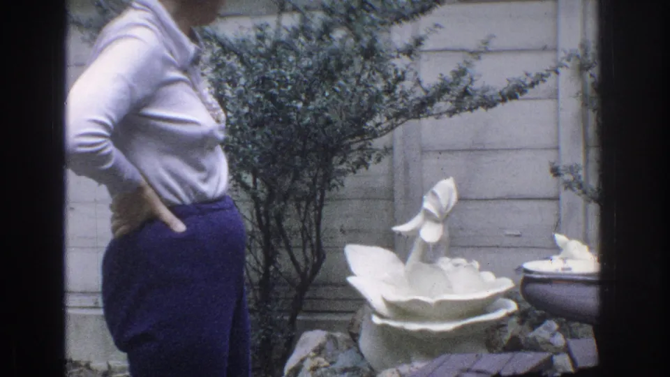 1971 Miami home-movie frame showing a woman in profile near a white garden fountain, shrubs, rocks, and a paneled fence.