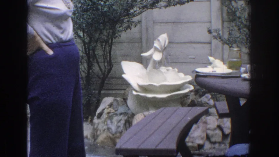 1971 Miami backyard frame with a white garden fountain, wooden bench or table, shrubs, rocks, fence, and part of a standing woman.
