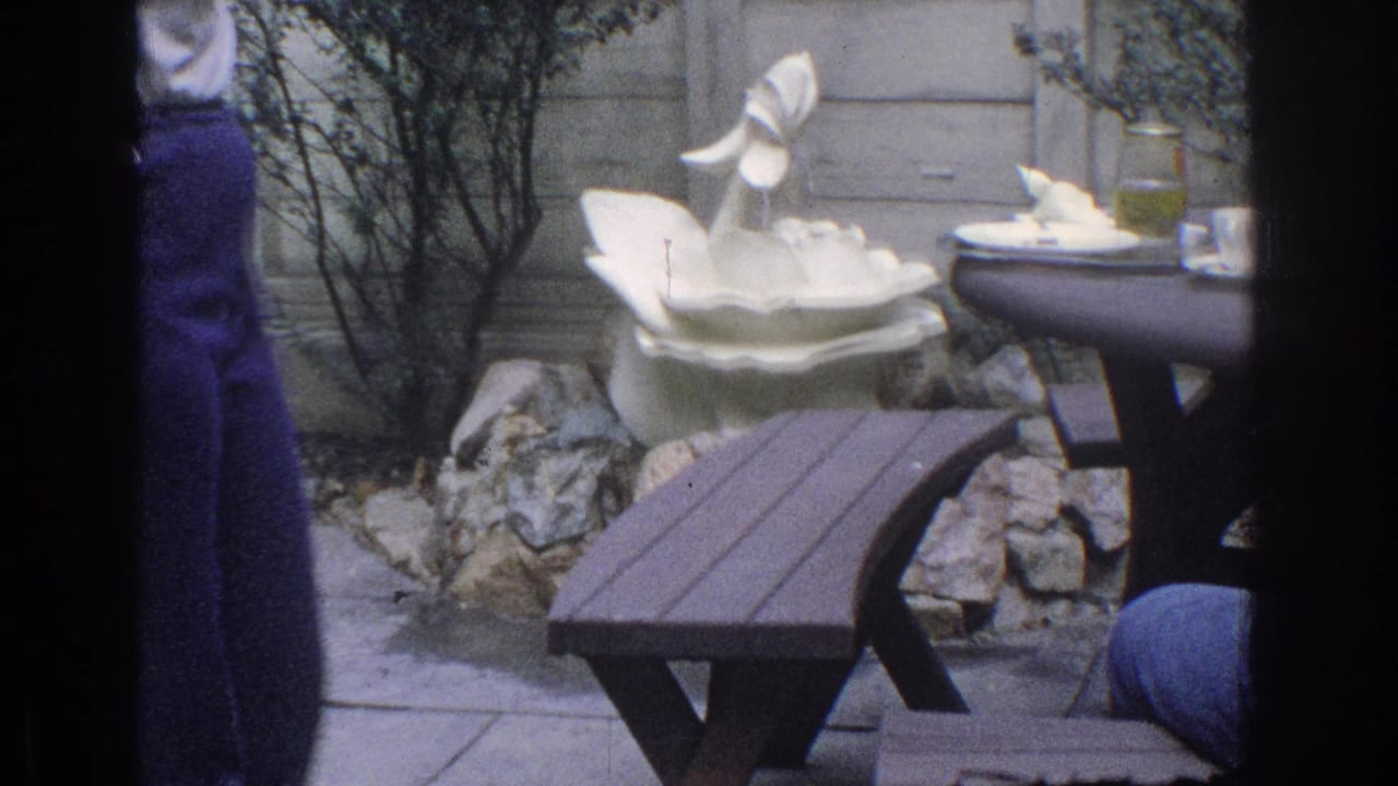 Garden patio table and bench beside a white ceramic swan fountain, with people partly visible at the edges.