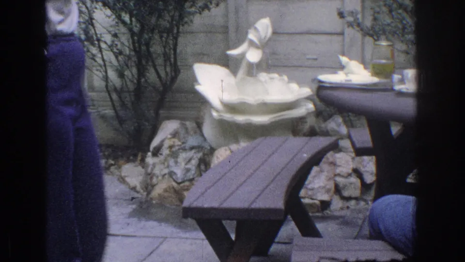 1971 Miami home-movie frame showing a garden patio table, bench, rock border, and white ceramic swan fountain with partial figures at the edges.