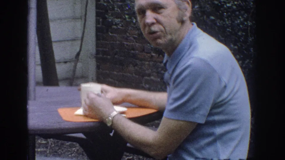 1971 Miami home-movie frame showing a man in a light blue polo holding a white cup at an outdoor garden table.