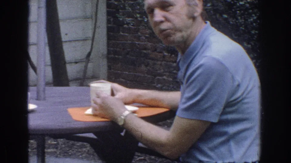 1971 Miami home-movie frame of a man seated at a garden table with a white cup, orange placemat, and breakfast items.