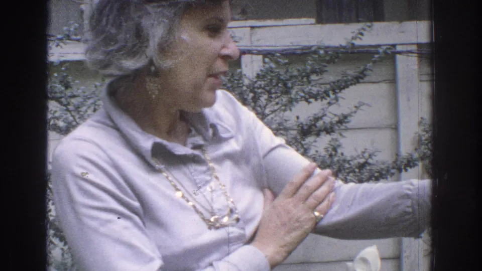 1971 Miami frame showing an older woman in profile beside a backyard table and shrubs.