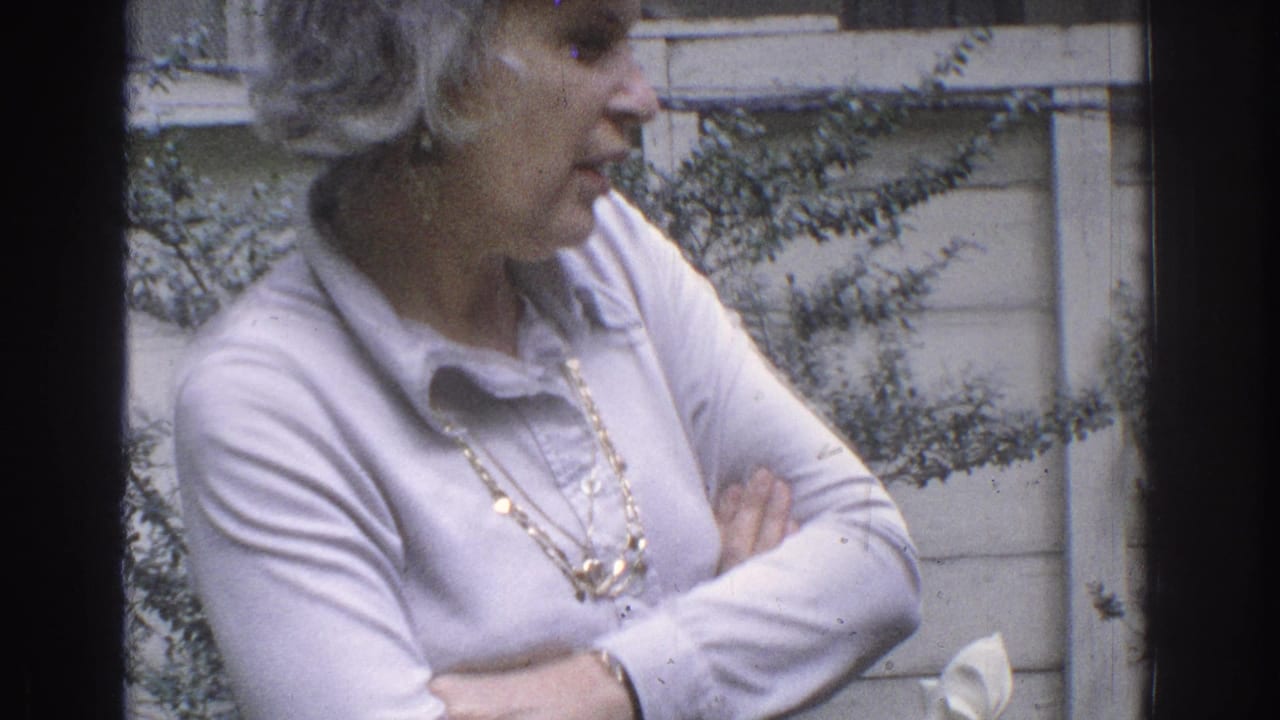 Woman with short gray hair stands outdoors with arms crossed near shrubs and wood siding.