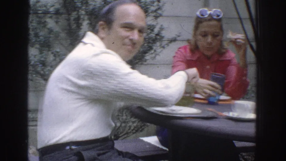 At an outdoor table in the supported Miami 1971 setting, a man in a white sweater turns toward the camera while a woman in red holds a cup.