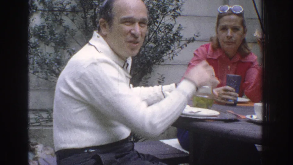 A man in a white sweater gestures during conversation at a patio table, while a woman in red sits behind cups and plates.