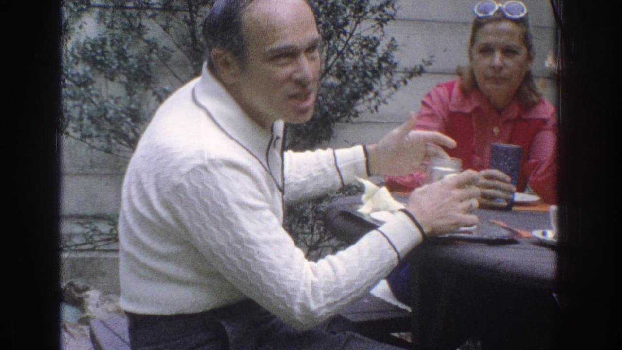 Man in a white sweater gestures at an outdoor table while a woman in a red shirt sits behind him.