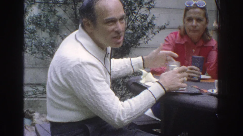 In supported 1971 Miami home-movie footage, a man in a white textured sweater gestures during an outdoor meal as a woman in red listens.