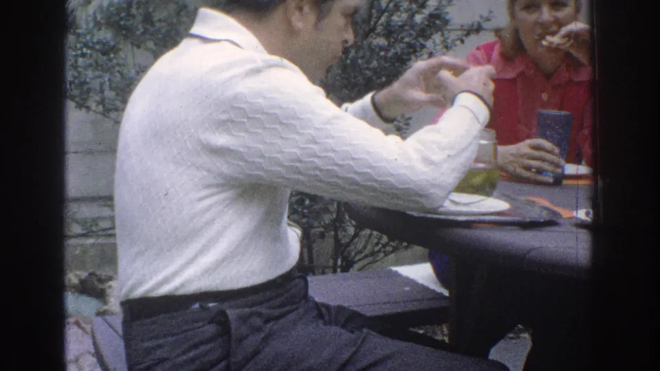 During the outdoor meal, the man leans forward with his hands raised while the woman in red eats at the table.