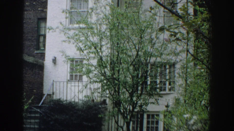1971 Miami home-movie frame showing lower windows, exterior railing, shrubs, and tree branches against a white brick facade.