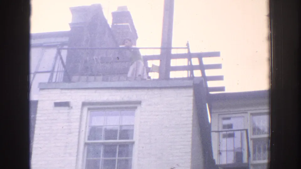 1971 Miami frame shifting upward to a rooftop with chimney forms, railing, pale sky, and a white brick wall with a window.