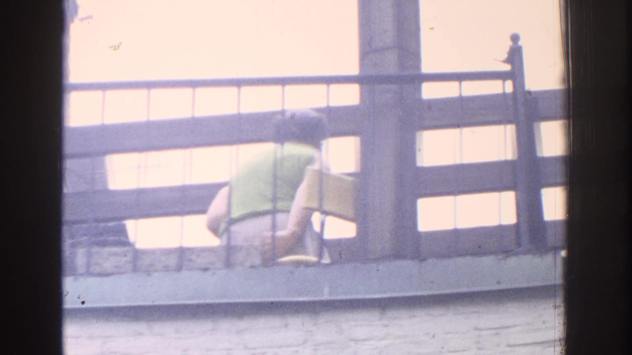 1971 Miami home-movie frame with a rear view of a woman in a green top sitting on a balcony beside a yellow object.