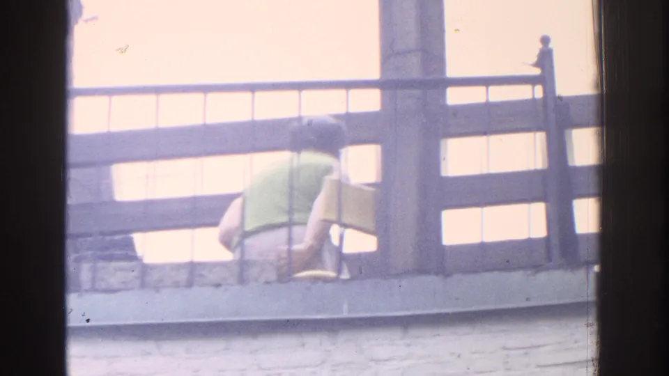 1971 Miami home-movie frame with a rear view of a woman in a green top sitting on a balcony beside a yellow object.