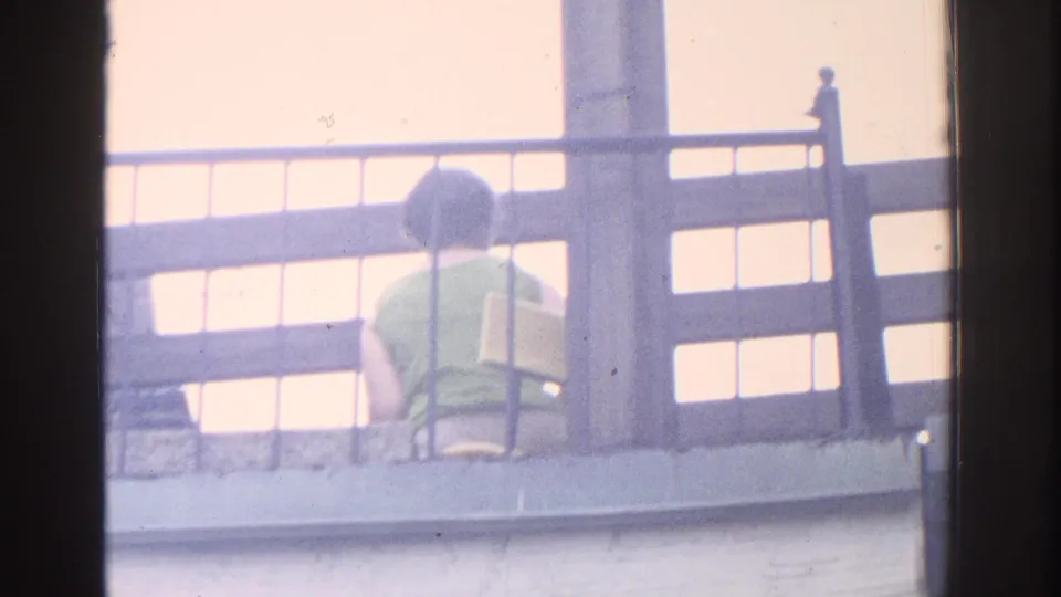 Soft 8mm frame from the supplied 1971 Miami clip showing the woman from behind on a balcony through dark foreground framing.