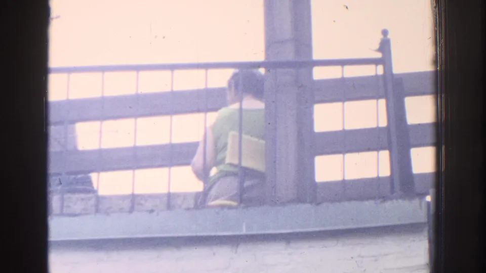 1971 Miami 8mm frame showing the woman seated near the balcony rail, slightly turned, with soft color and window-framed edges.