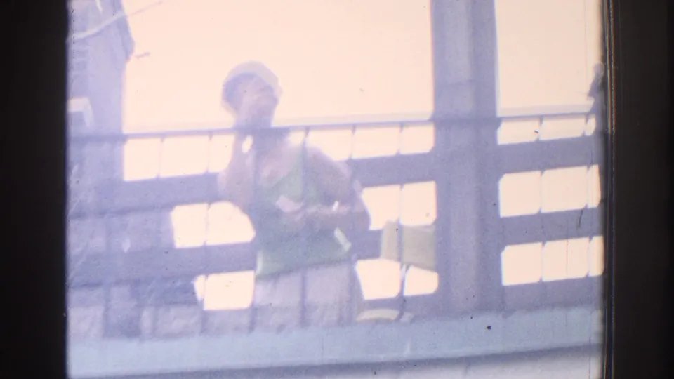 1971 Miami home-movie frame showing a woman on a balcony, standing beside railings in washed-out exterior light.