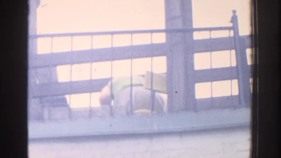 1971 Miami home-movie frame with the balcony railing dominating as the woman moves low or bends behind it.