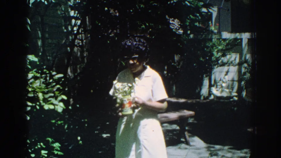 1971 Miami home-movie frame showing a woman in white holding a potted plant amid dense garden foliage.