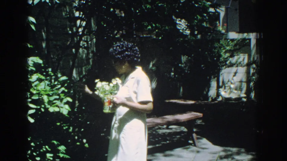In 1971 Miami home-movie footage, a woman in a white dress stands among dense garden foliage holding a small flower arrangement.