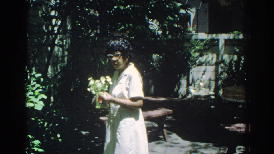 A woman in a white dress turns slightly toward the camera while holding flowers in a sunny Miami garden setting.