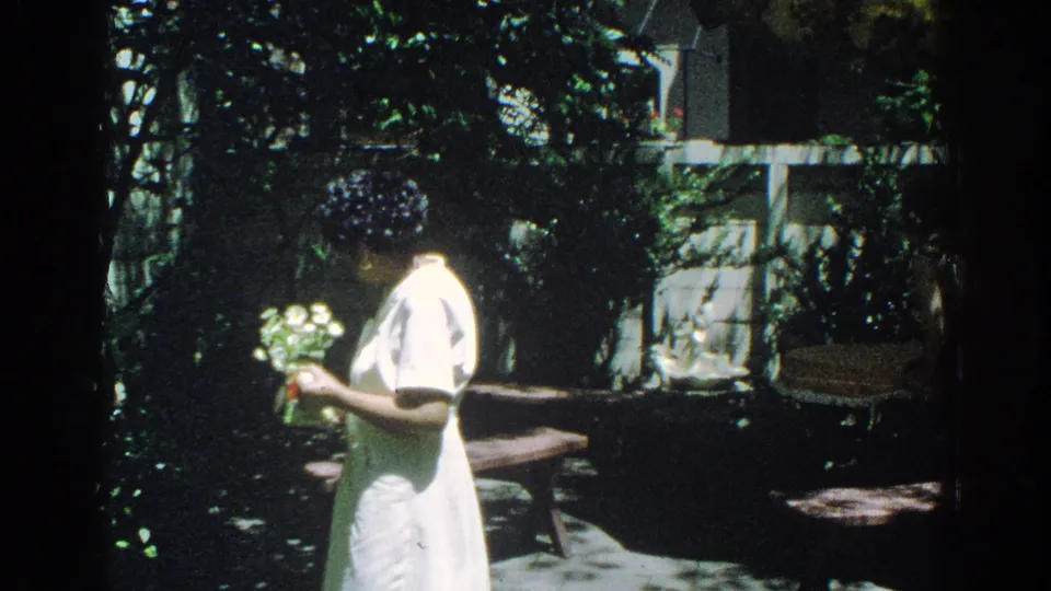 The woman looks down at her flowers in the garden, framed by deep shade, sunlit leaves, and patio furniture.