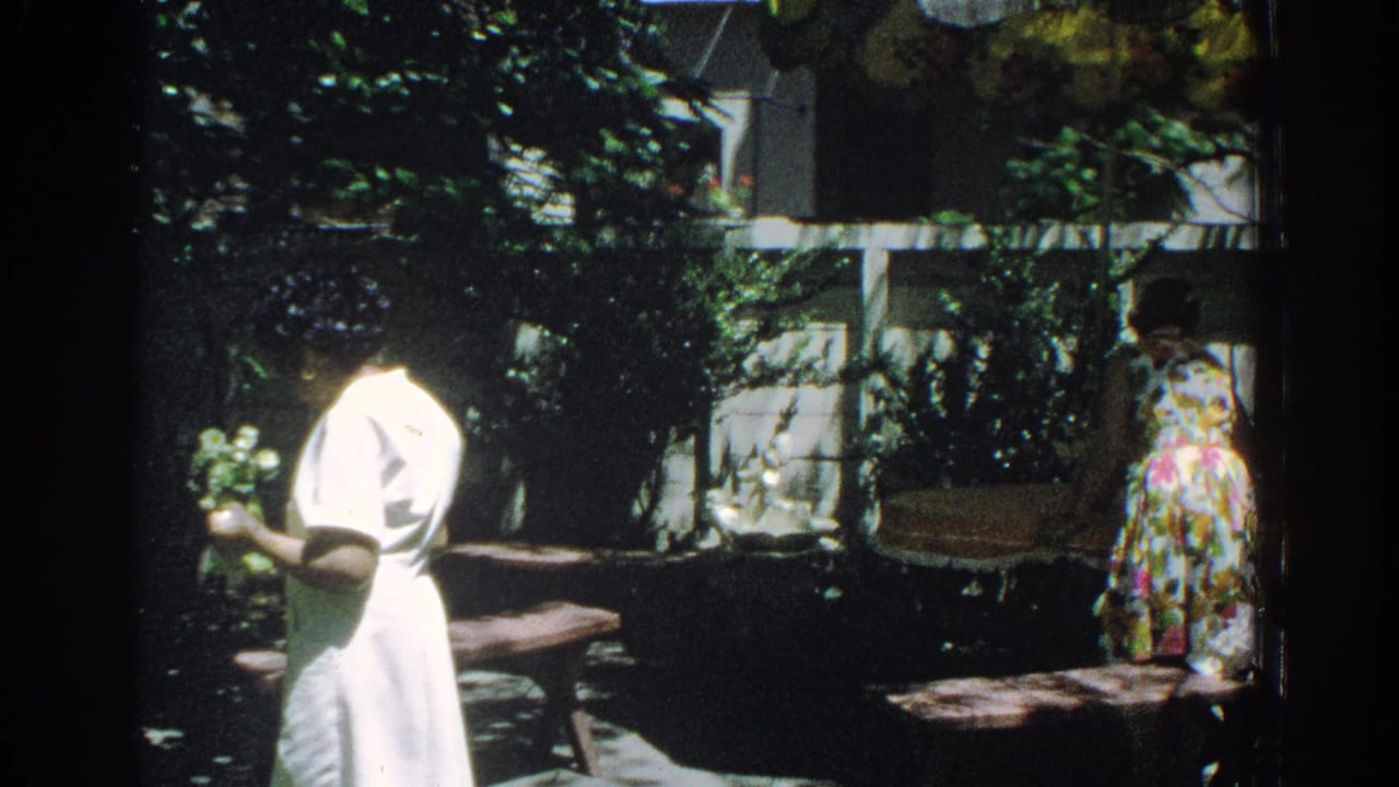 1971 Miami backyard frame with a woman holding flowers in the foreground and a second woman in a floral dress near the fence.
