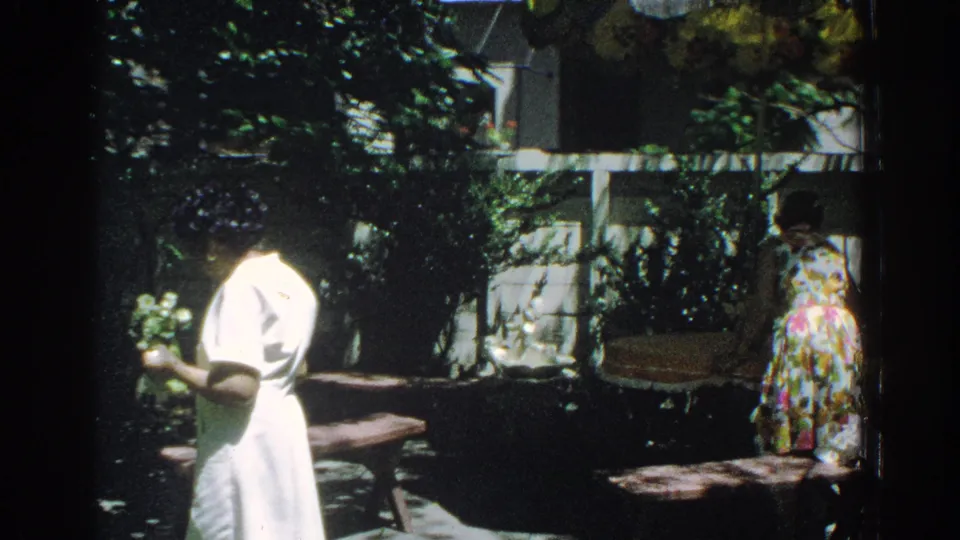 1971 Miami backyard frame with a woman holding flowers in the foreground and a second woman in a floral dress near the fence.