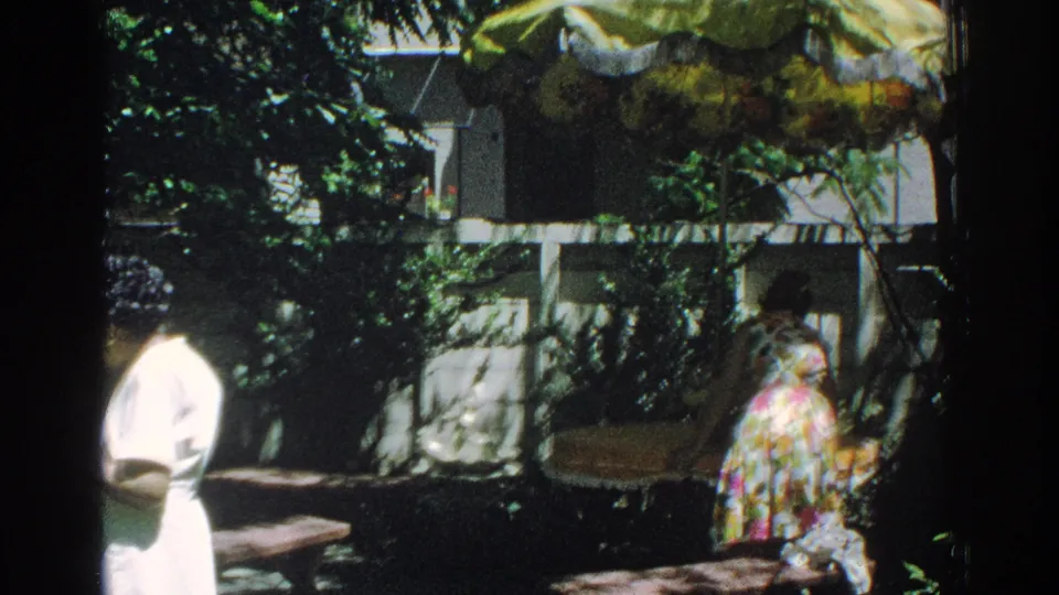 1971 Miami home-movie frame with one woman in white at the edge of the shot and another in a floral dress by benches and foliage.
