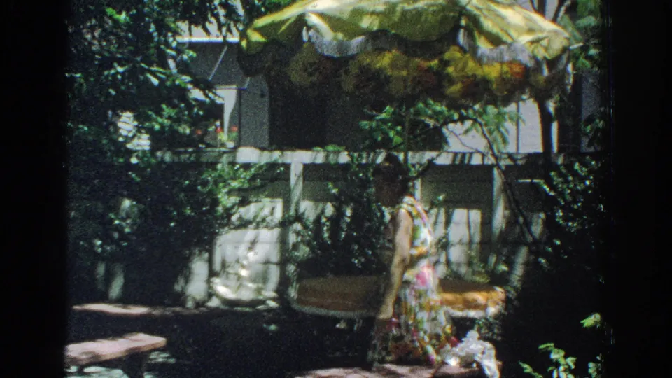 1971 Miami frame showing a woman in a colorful floral dress moving through a backyard with benches, plants, and a white fence.