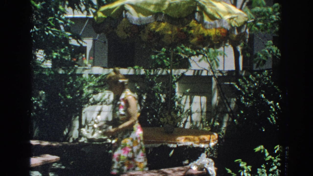 Woman in a floral dress beside a patio table under a sunshade in a leafy backyard.