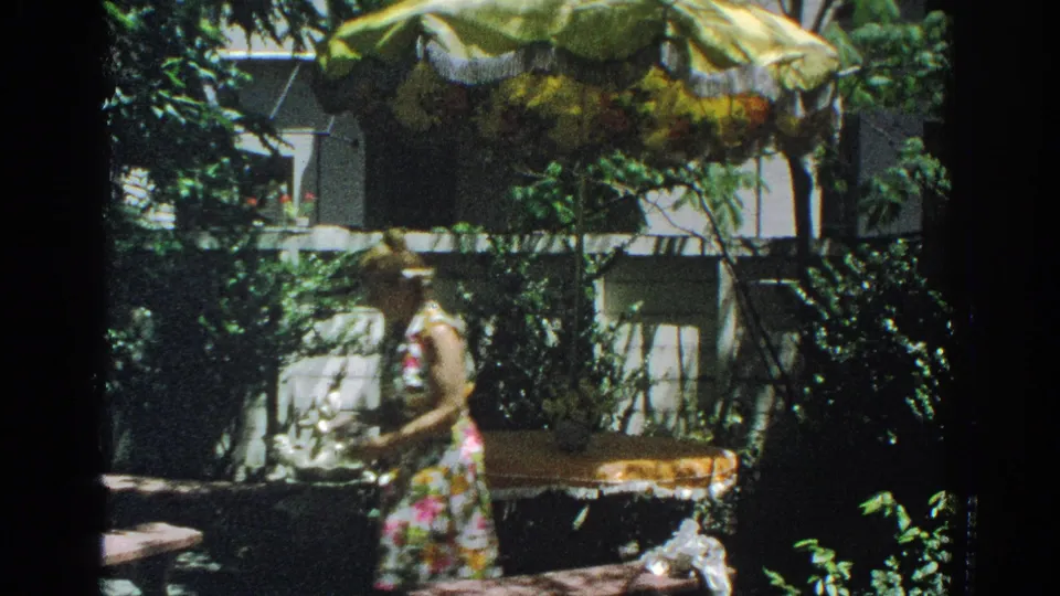 1971 Miami 8mm frame showing a woman in a floral dress near a round patio table, sunshade, white fence, and garden foliage.