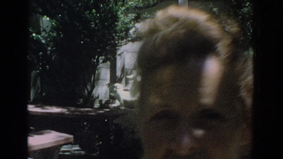 1971 Miami 8mm frame with a soft-focus close view of a woman's face in front of a shaded backyard, foliage, and patio table.