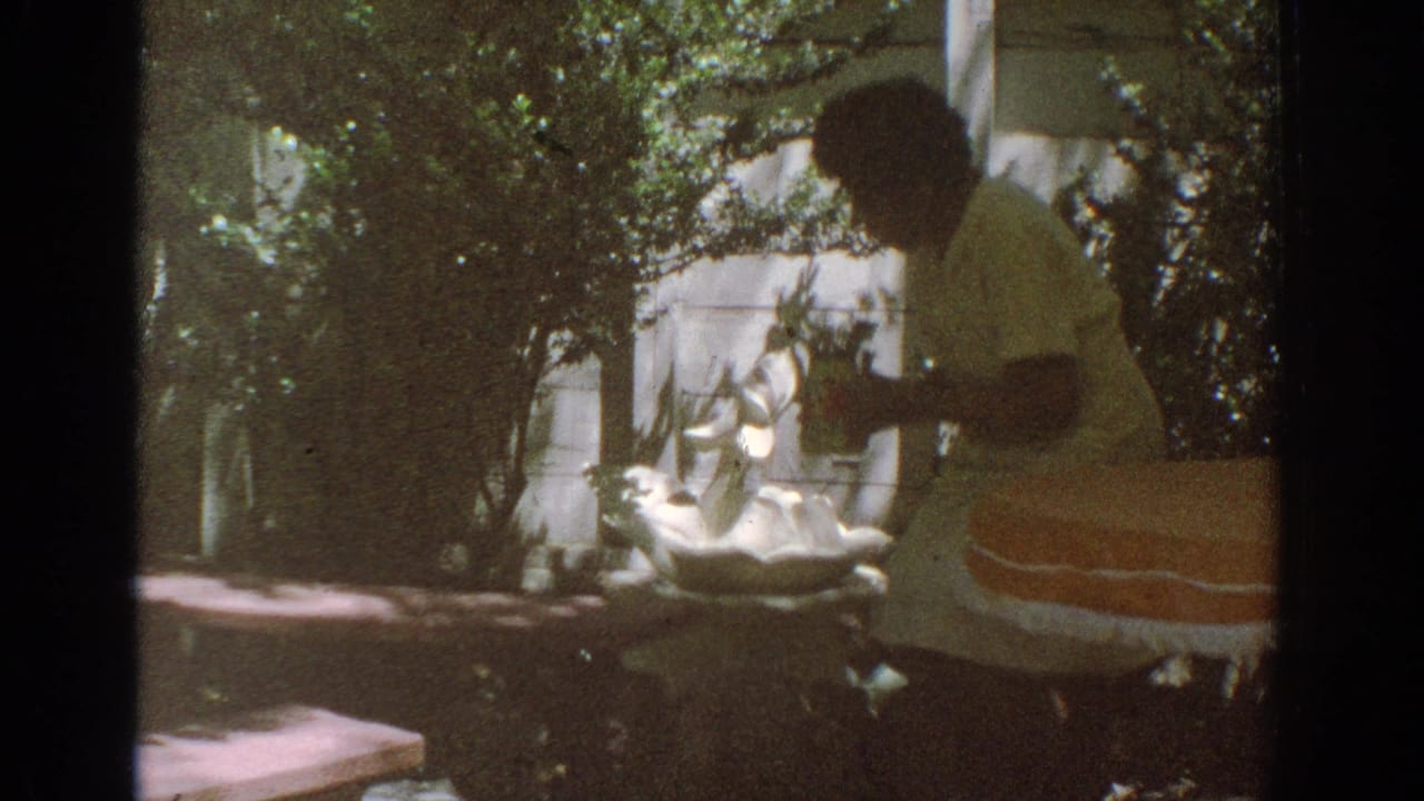 In the 1971 Miami garden footage, a woman in a light dress tends a white decorative fountain amid leafy shade.