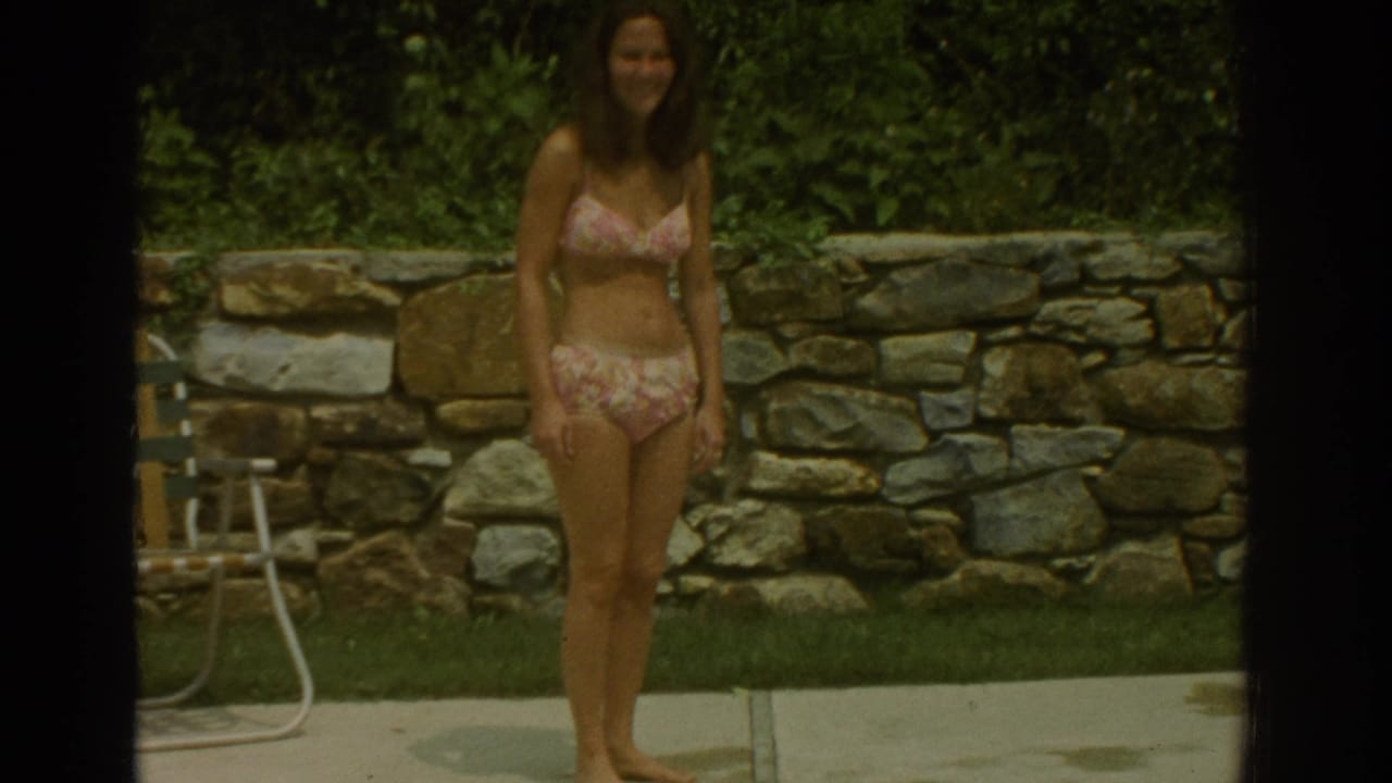 Woman in a pink floral bikini stands barefoot near a backyard pool and stone wall.
