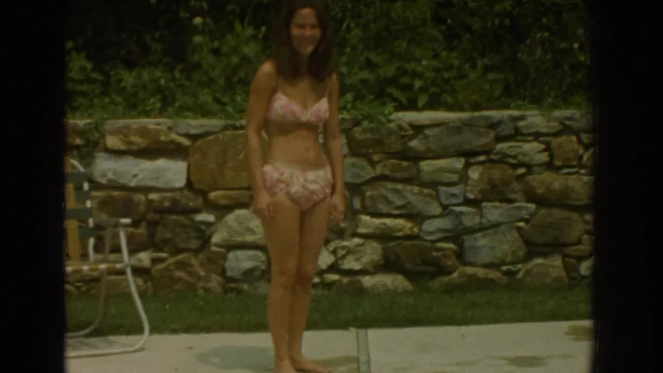 In 1971 Miami home-movie footage, a woman in a pink floral bikini stands on a concrete pool deck near a stone wall and greenery.
