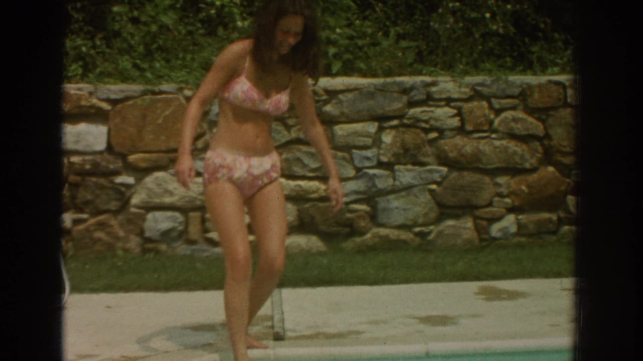 The woman walks barefoot along the concrete pool edge in 1971 Miami, with a stone wall and greenery behind her.