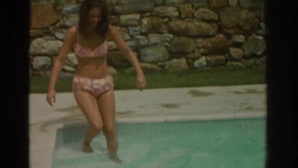 The woman steps down into the backyard pool, partly in the water, with the stone wall and concrete deck visible behind her.