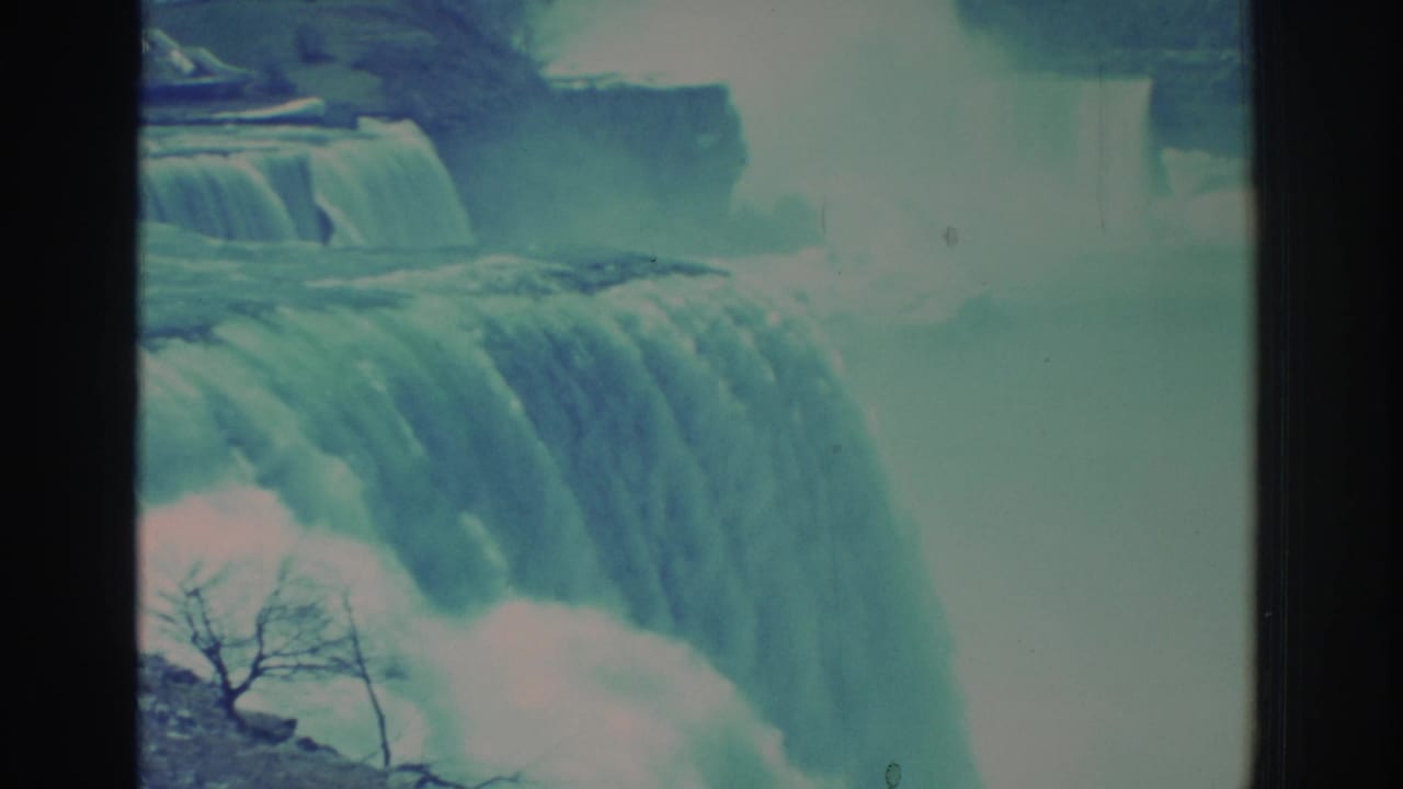 1971 8mm frame of Niagara Falls from a close visitor angle, showing water cascading over the edge, mist, and a bare branch in the foreground.