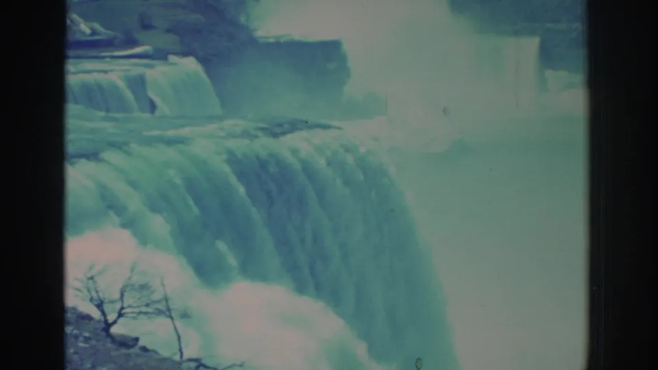 1971 8mm frame of Niagara Falls from a close visitor angle, showing water cascading over the edge, mist, and a bare branch in the foreground.