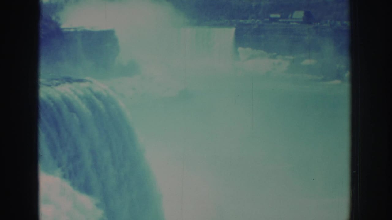 1971 Niagara Falls 8mm view with a curtain of water dropping into mist, distant shoreline detail, and dark film border at the side.