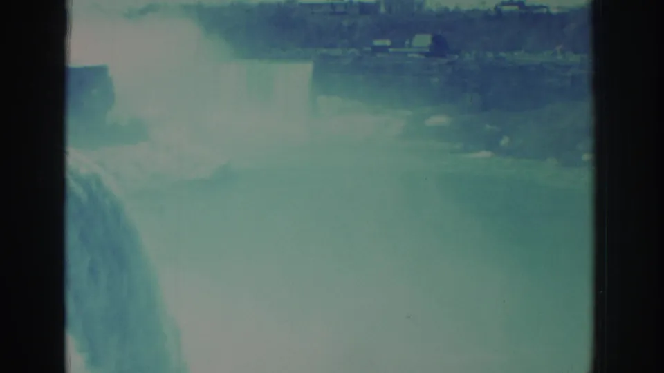 1971 8mm Niagara Falls frame with mist dominating the lower view, water dropping at left, and small distant structures near the shoreline.