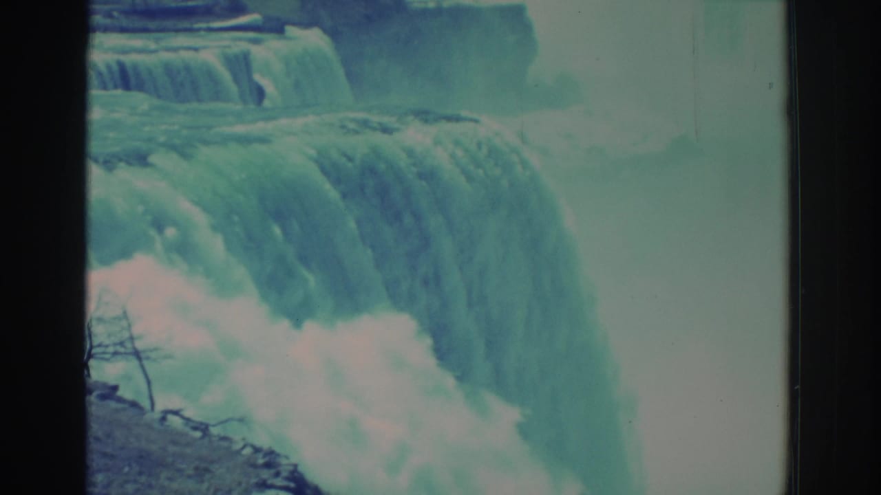 1971 8mm home-movie view of Niagara Falls with cascading water, mist, rocky edges, teal color cast, and visible frame borders.
