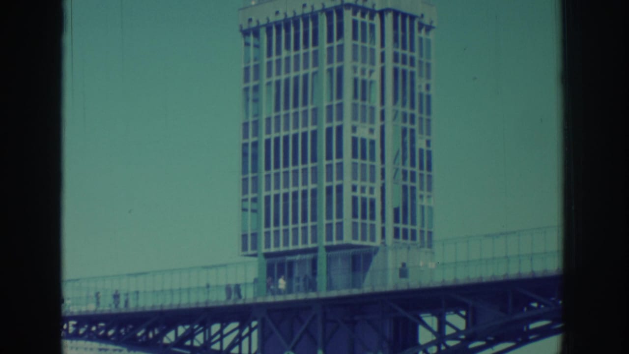 1971 Niagara Falls home-movie frame showing a gridded tower over a bridge, with tiny pedestrians and faded teal sky.