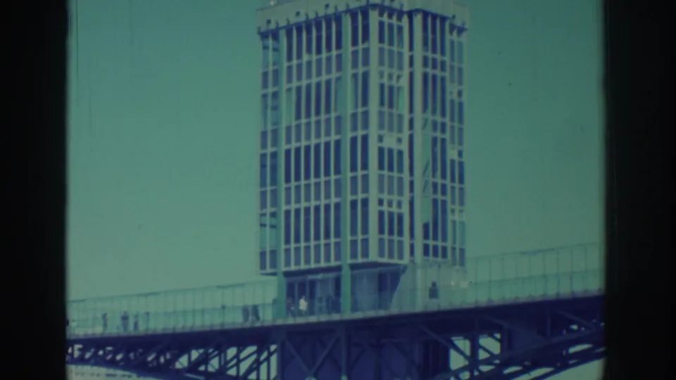 1971 Niagara Falls home-movie frame showing a gridded tower over a bridge, with tiny pedestrians and faded teal sky.
