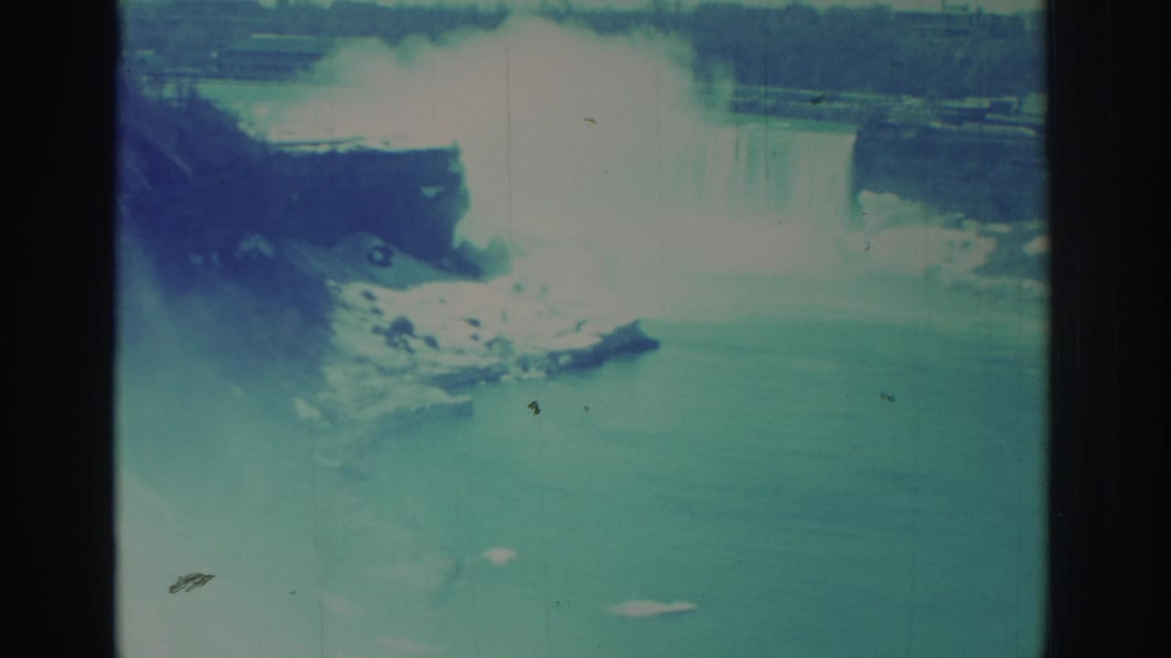 Distant Niagara Falls view in archival 8mm color, with a rocky point, river water, and dense mist from the cascade.