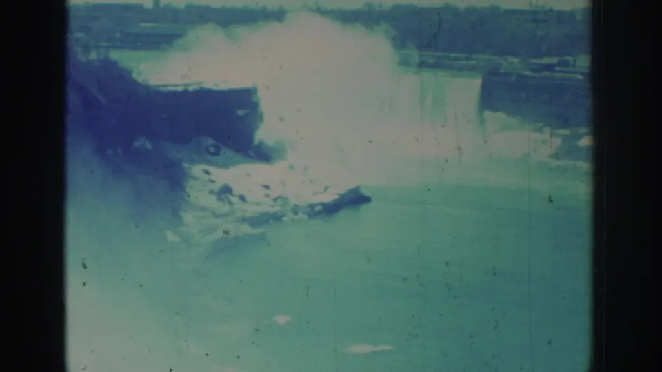 1971 Niagara Falls home-movie frame showing river water, a rocky shoreline, and a misty waterfall in the background.