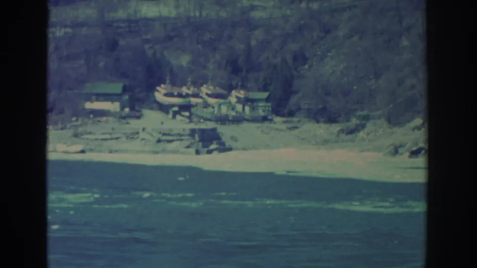 Vintage 8mm frame of a Niagara Falls-area shoreline with boats, low structures, water, and a steep wooded bank.