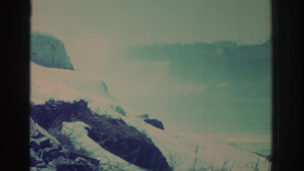 1971 8mm Niagara Falls area view with snow-covered rock, misty water, bare brush, and faded blue-green film color.