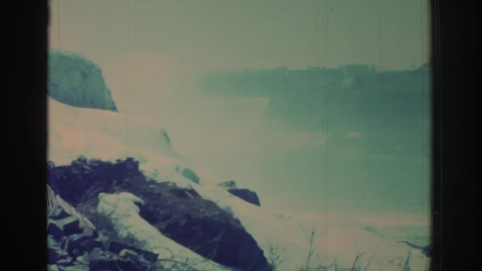 1971 8mm Niagara Falls area view with snow-covered rock, misty water, bare brush, and faded blue-green film color.