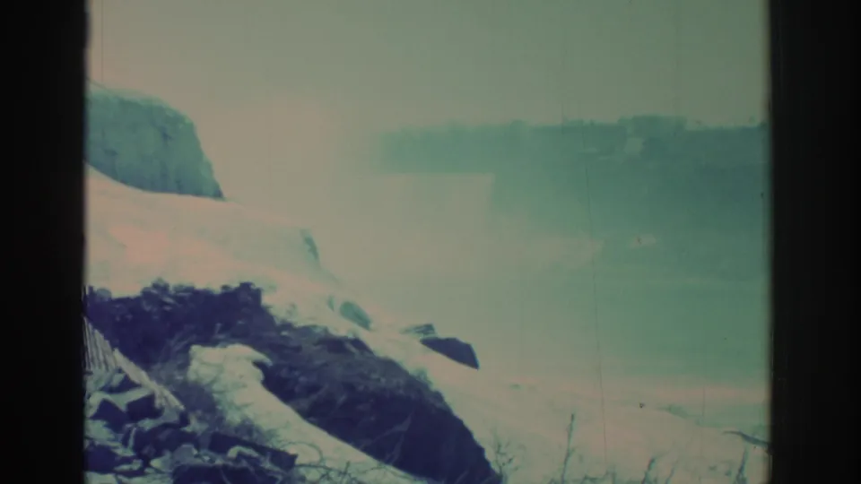 Faded 8mm frame of the Niagara Falls area, showing snow, dark rock, mist over water, and a heavy black film border.